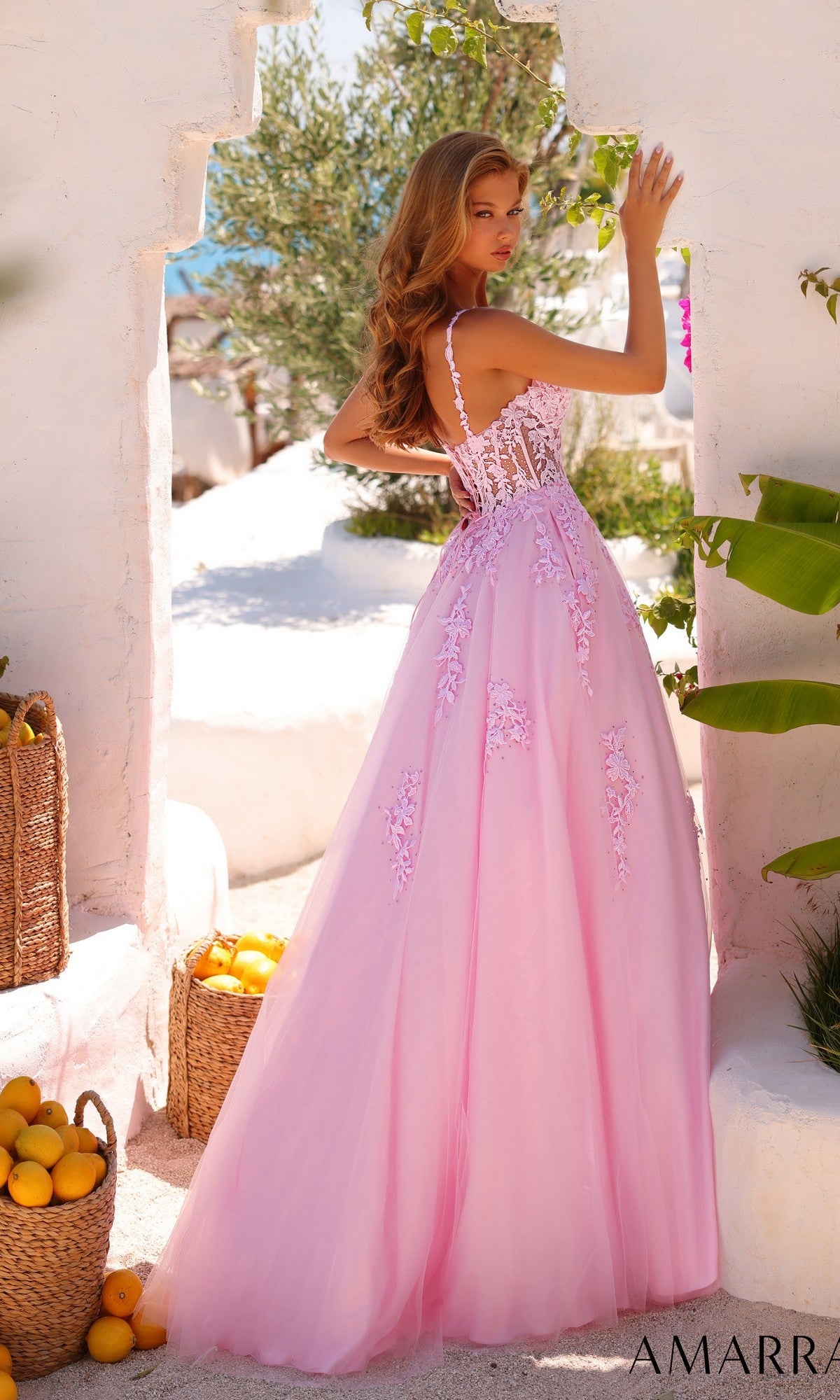 A woman stands outdoors near white walls and baskets of yellow fruit, wearing the Sheer-Waist Long A-Line Prom Dress: Amarra 89523 in light pink, with greenery in the background.